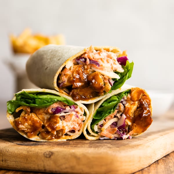 Three BBQ chicken wraps with coleslaw and lettuce, stacked on a wooden board.