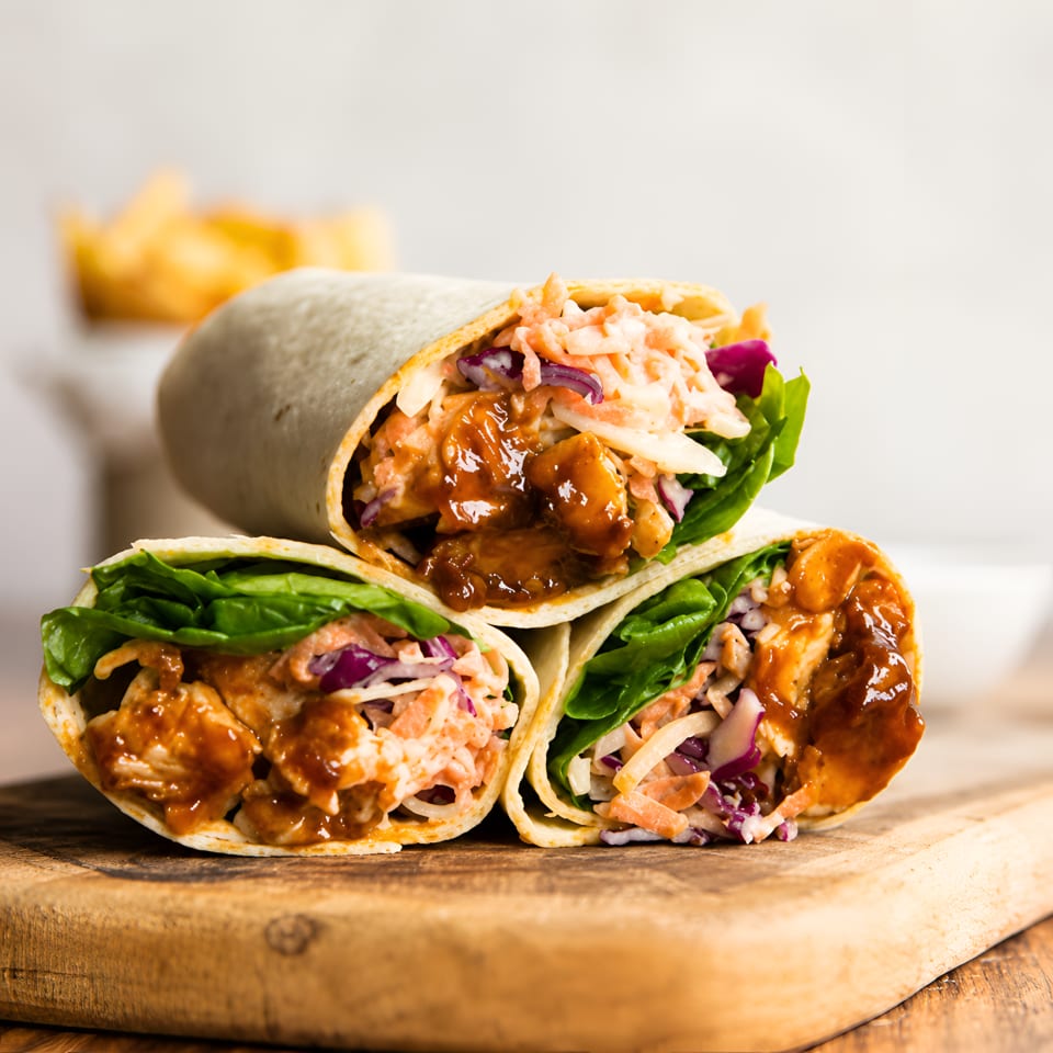 Three BBQ chicken wraps with coleslaw and lettuce, stacked on a wooden board.