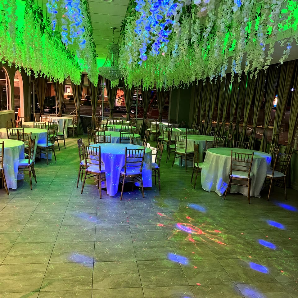 Restaurant dining room with tables set, illuminated by green and blue lights, and hanging floral decor.