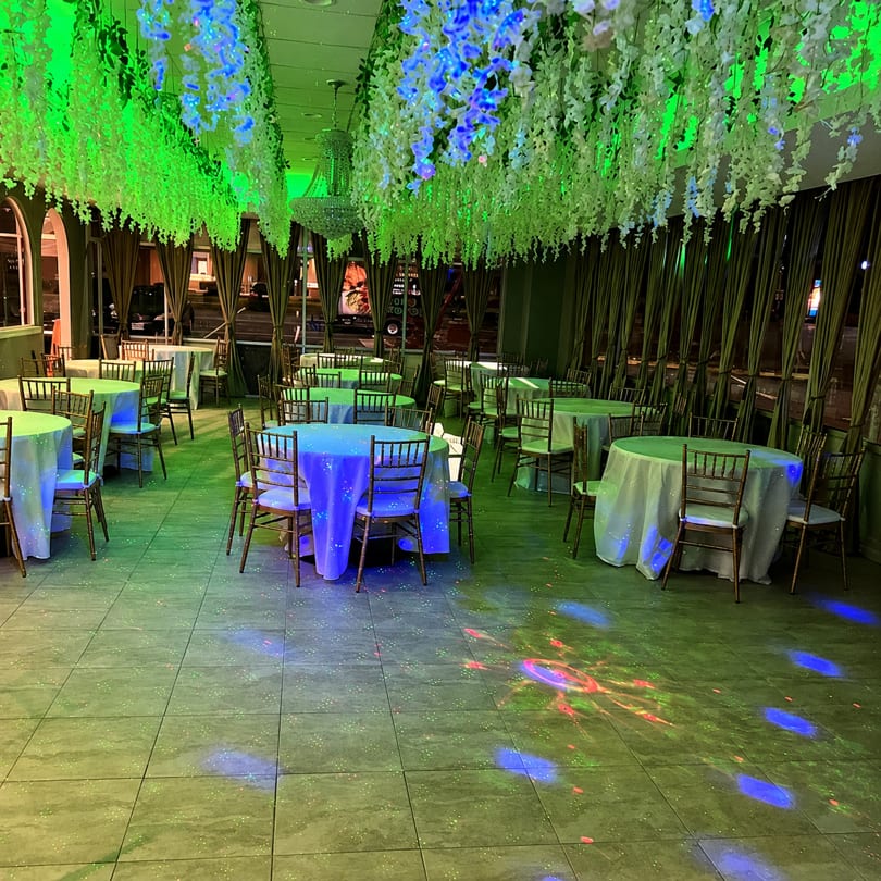 Restaurant dining room with tables set, illuminated by green and blue lights, and hanging floral decor.