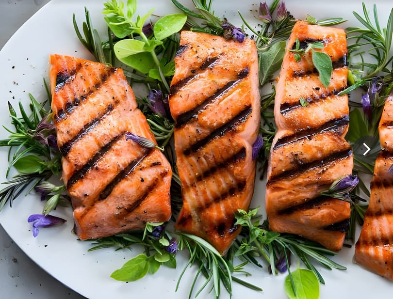 Grilled salmon fillets with grill marks, garnished with fresh rosemary, herbs, and purple flowers.