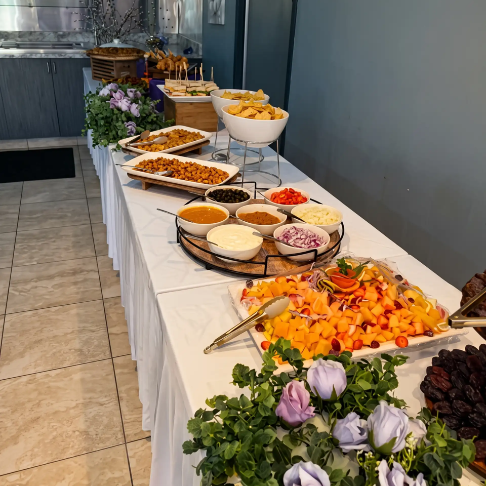 Elegant buffet table displaying appetizers, chips, dips, fruit, olives, sandwiches, and decorative flowers in a modern dining space.