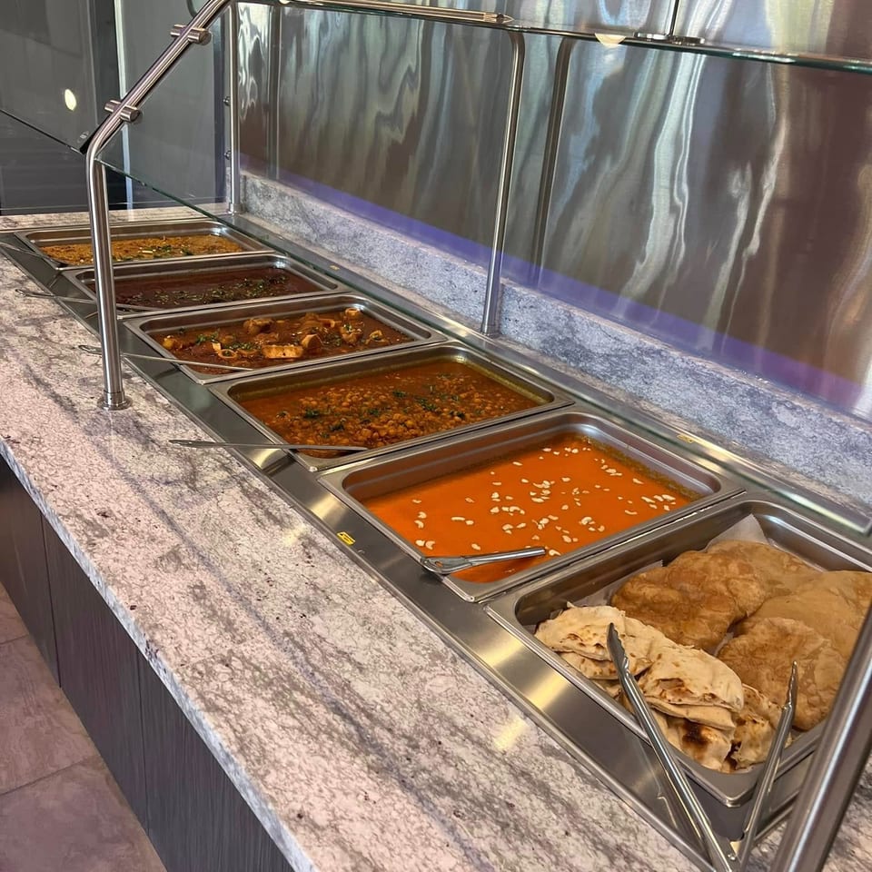 Buffet counter displaying trays of curries, lentils, stewed dishes, and naan beneath a glass sneeze guard.