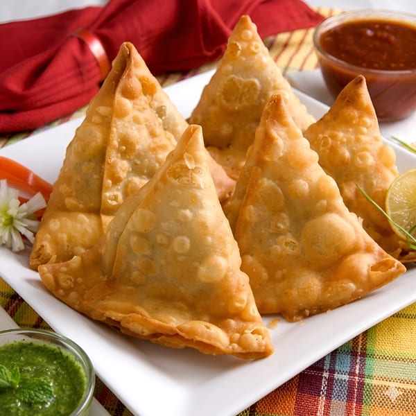 Golden-fried samosas on a white plate with green and red chutneys, garnished with lemon and herbs.