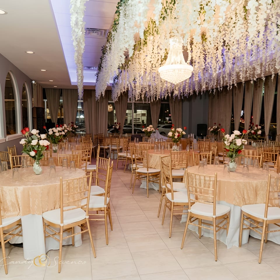Elegant banquet hall with gold-draped tables, gold chairs, floral centerpieces, and hanging white wisteria.